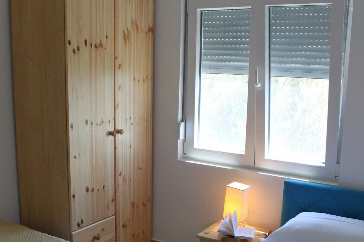 A bedroom with two beds, a wooden wardrobe, a bedside table with a lamp, and a window with blinds.