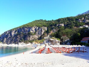 A beach with white sand, red lounge chairs, and umbrellas, with a rocky cliff and greenery in the background. - Blue Mediterranean (Petrovac)