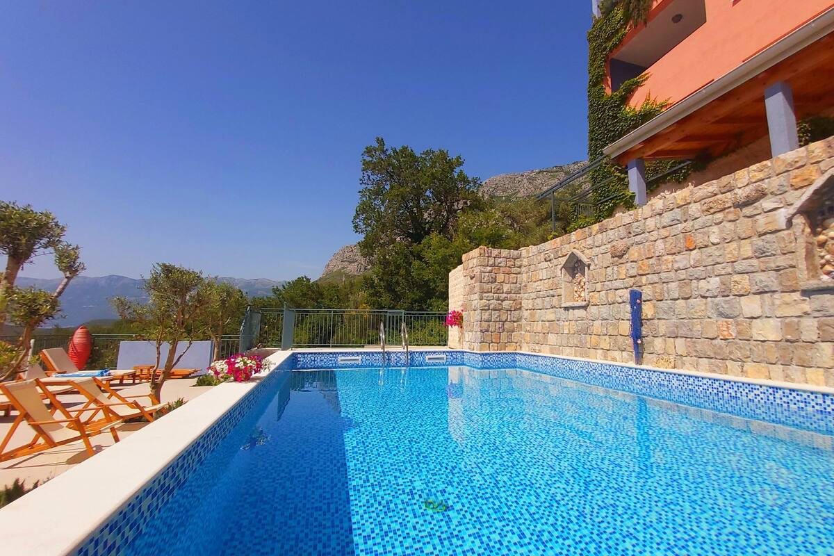 A swimming pool with blue mosaic tiles, surrounded by lounge chairs and a stone wall with decorative elements.