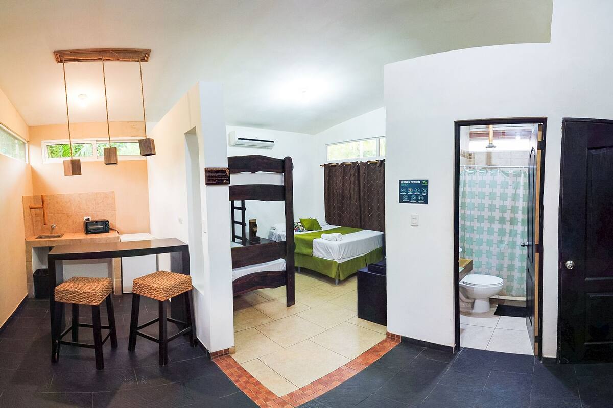 A compact hotel room with a kitchenette, a bed with green bedding, a wooden bunk bed, a shower area, and a small sitting area with stools.