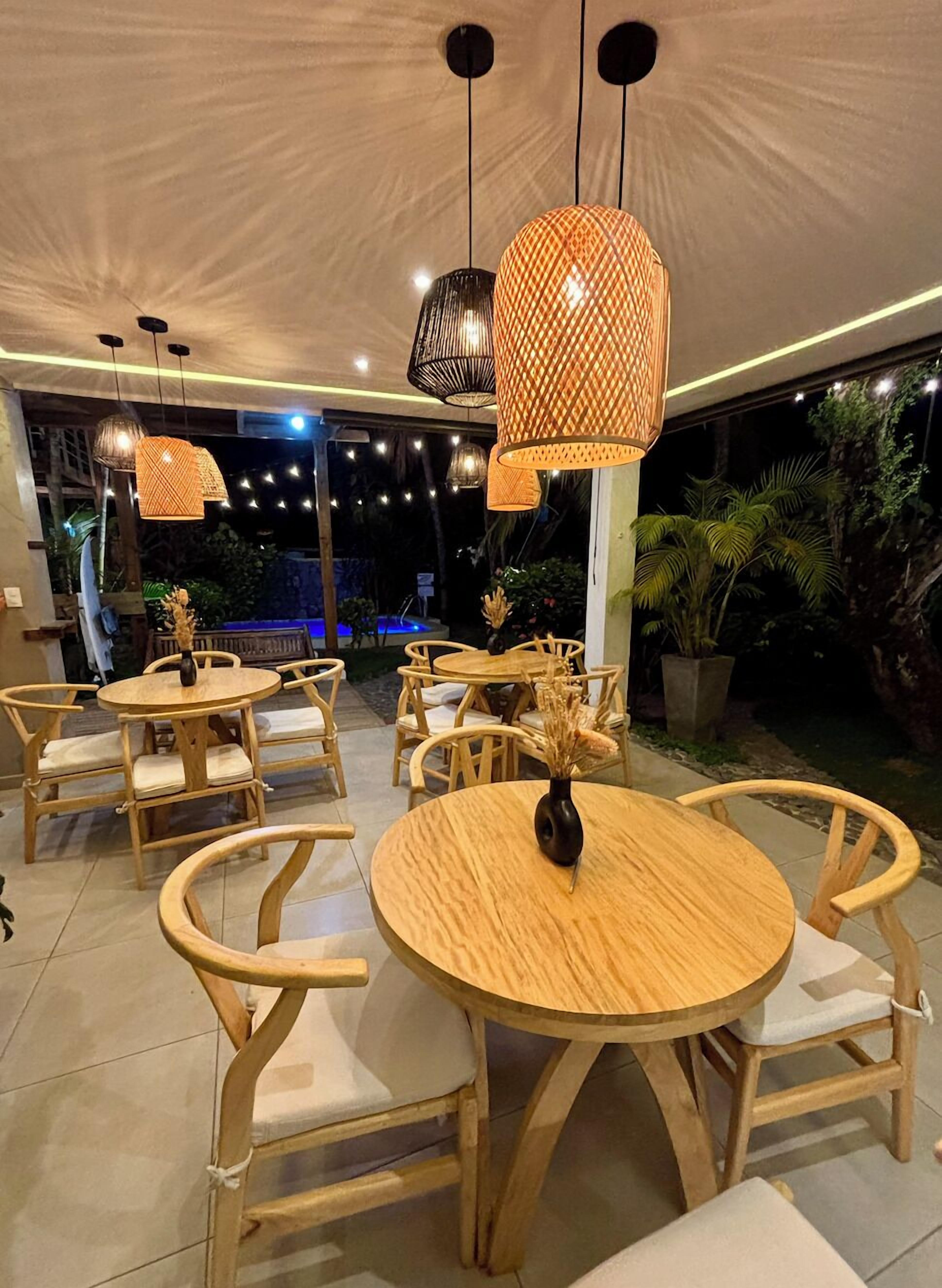 Un restaurante con mesas y sillas de madera redondas, lámparas colgantes de ratán y vistas a una piscina y follaje exterior.