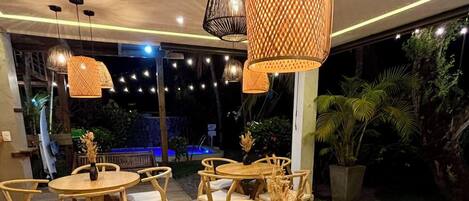 Un restaurante con mesas y sillas de madera redondas, lámparas colgantes de ratán y vistas a una piscina y follaje exterior.