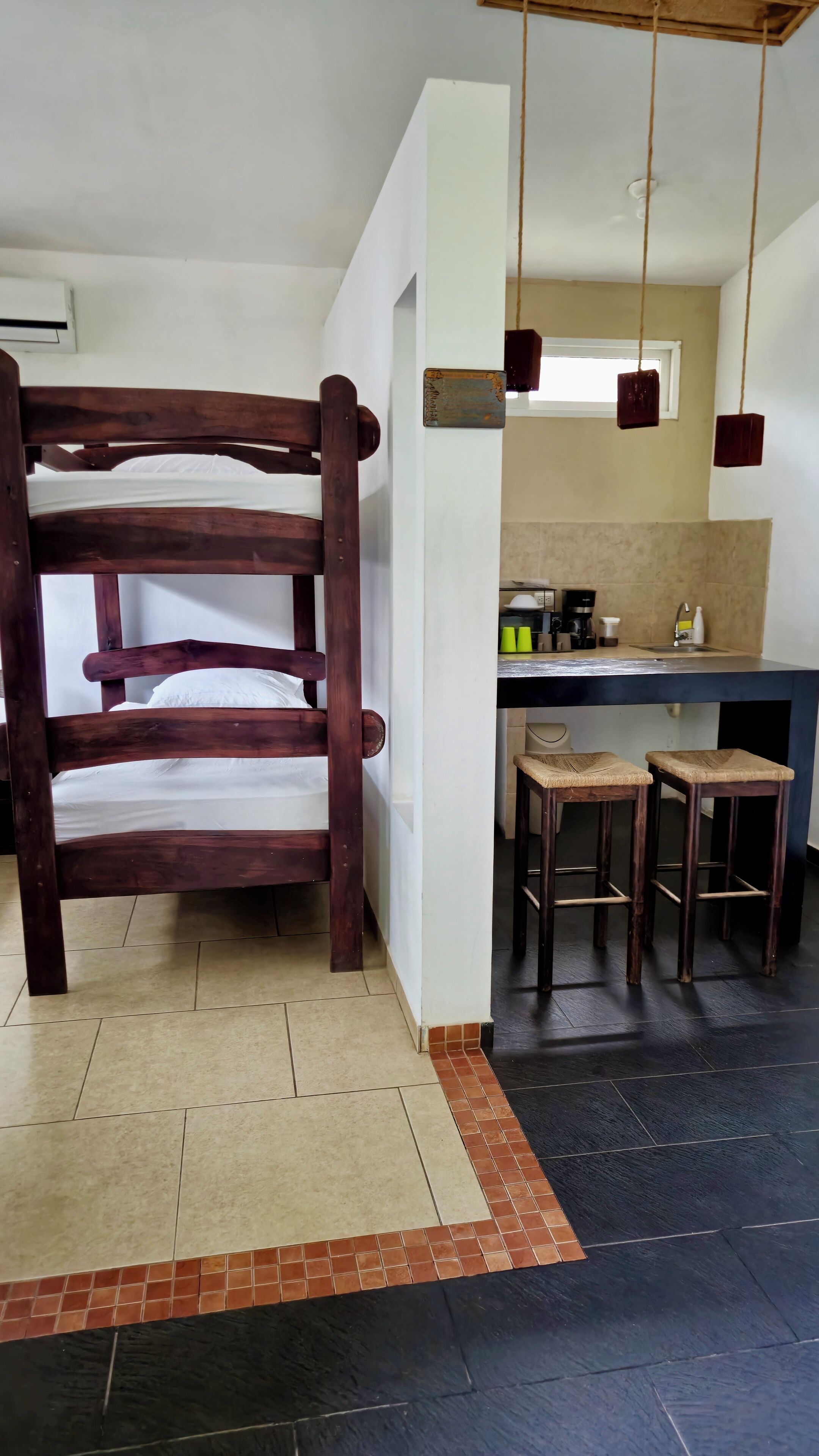 Une pièce avec un lit superposé en bois, un coin cuisine avec des tabourets et un sol carrelé.