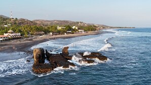 Paisaje costero con formaciones rocosas en el agua, una playa con gente y edificios al fondo.