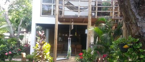 Un edificio de dos pisos con balcón, rodeado de vegetación exuberante y una hamaca.