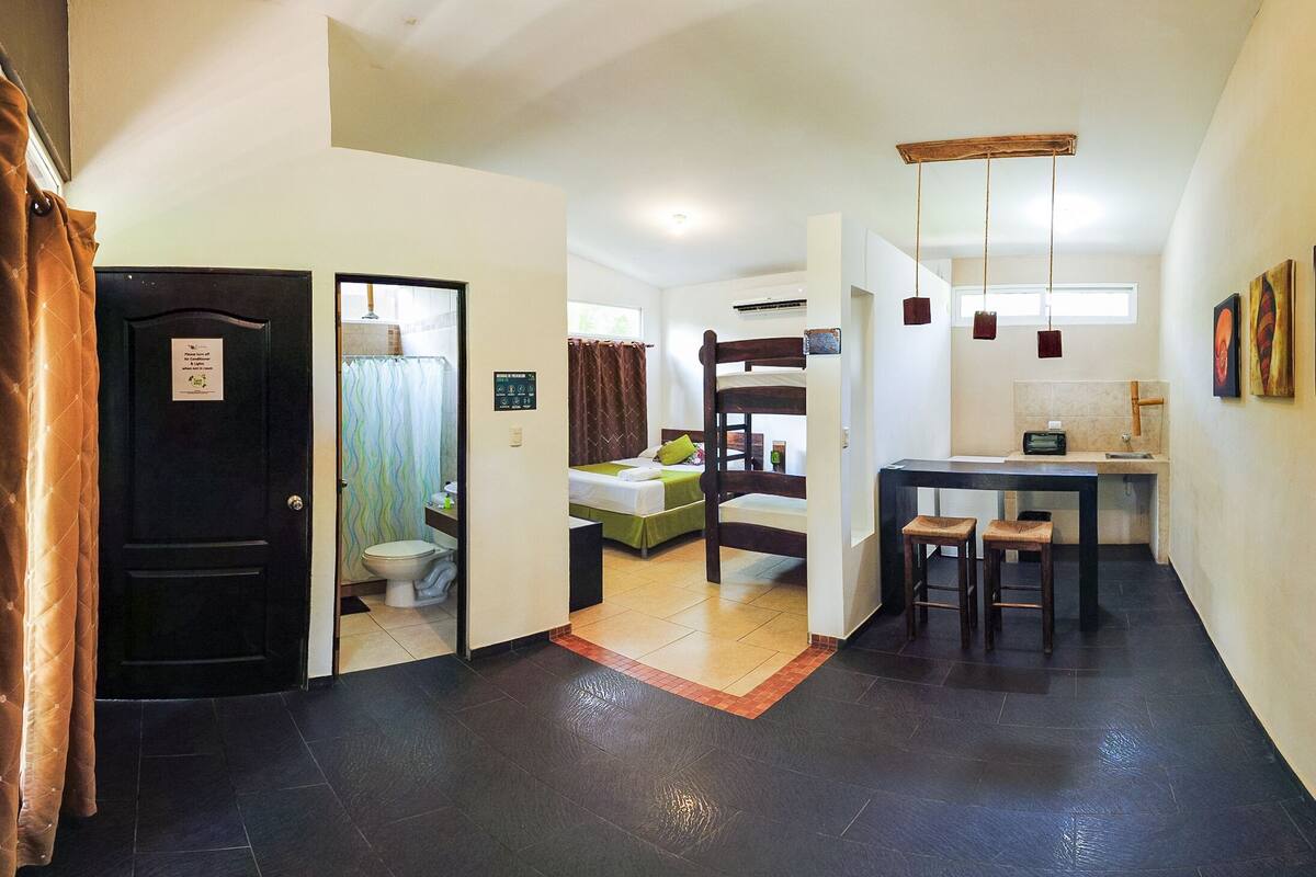 A compact hotel room with a bathroom, a bed, a small kitchenette, and a dining area.