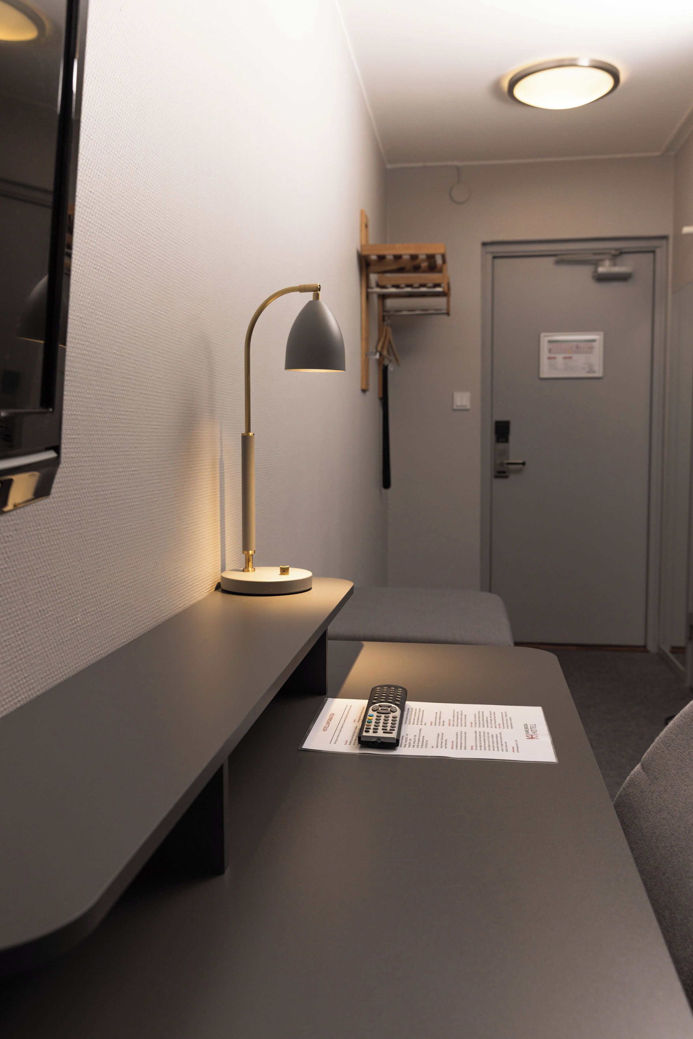Ett hotellrum med ett skrivbord, en lampa, en fjärrkontroll och en dörr.