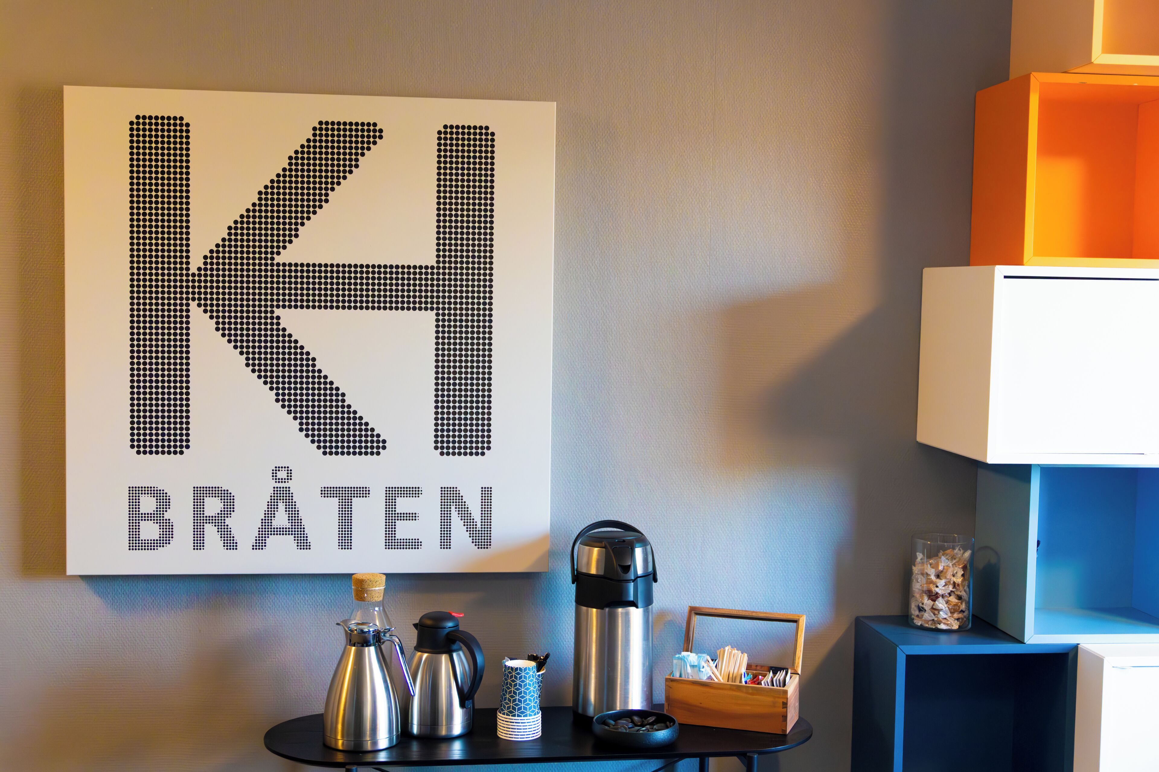 A wall with a large letter 'K' and the word 'BRÄTEN' in a room with a coffee maker, thermos, and a basket of tissues.