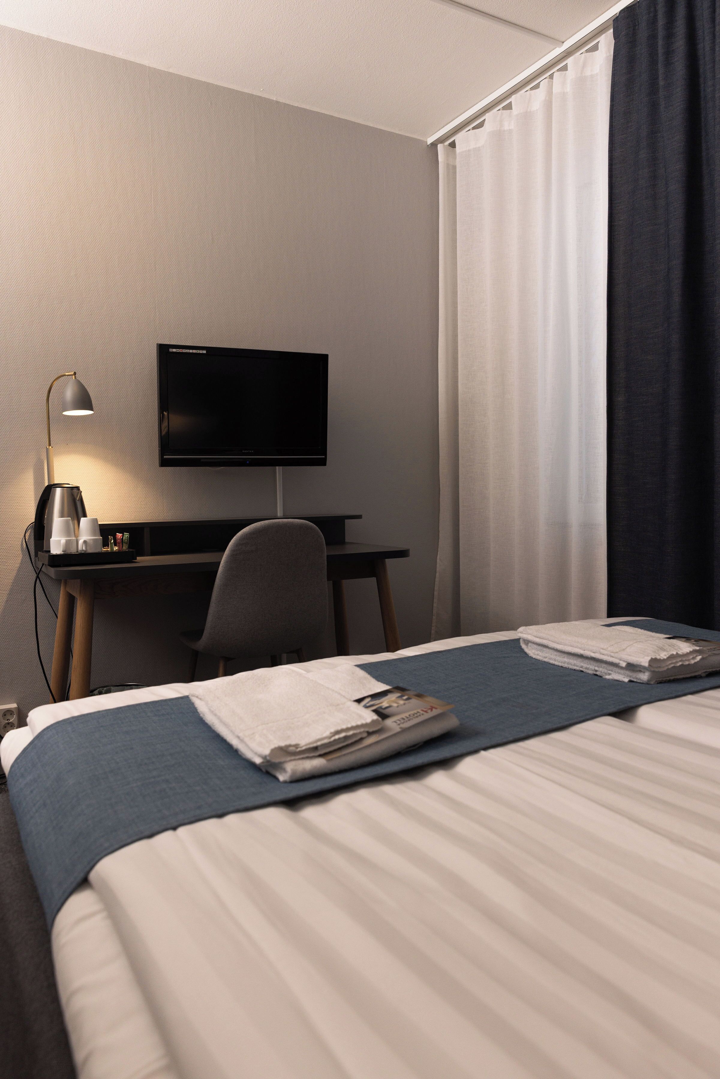 Habitación de hotel con cama, escritorio con computadora y lámpara, televisor y ventana con cortinas.