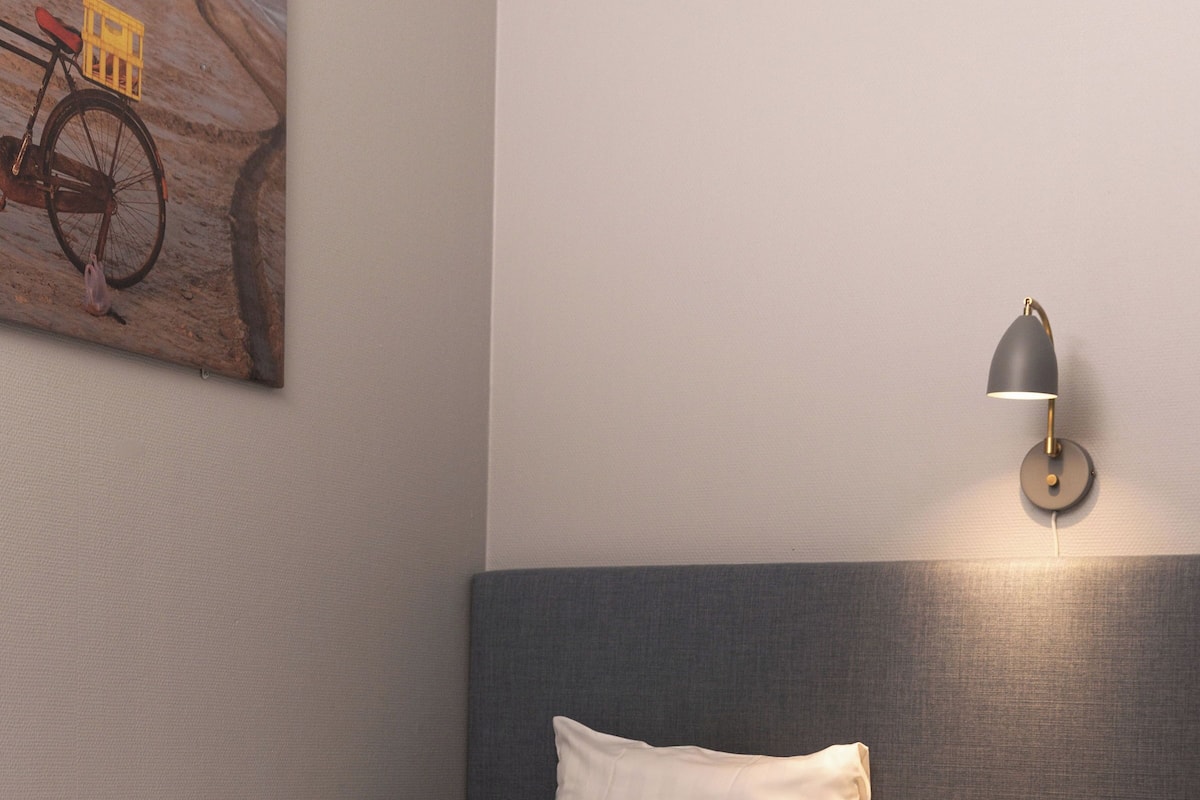 A neatly made bed with a gray headboard, a small wooden stool, and a wall-mounted lamp.