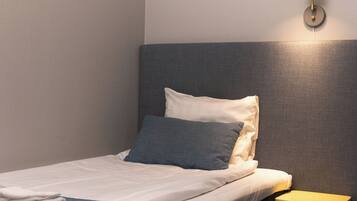 A neatly made bed with a gray headboard, a small wooden stool, and a wall-mounted lamp.