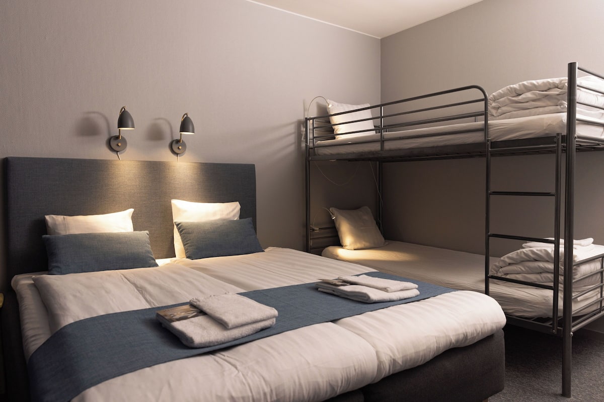 A hotel room with a large bed, a smaller bed, and a bunk bed. There are wall-mounted lights and a headboard.