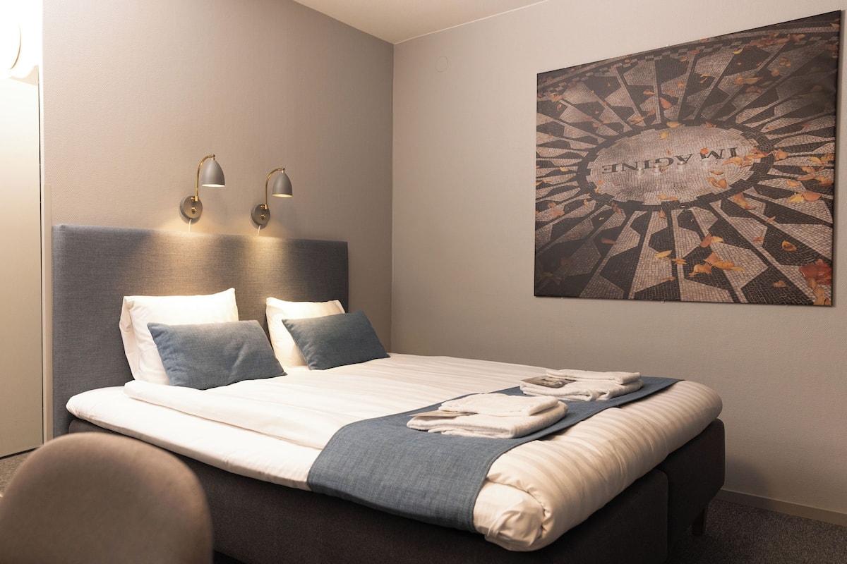 A neatly made bed with a blue blanket and pillows, two wall-mounted lamps, and a framed artwork on the wall.