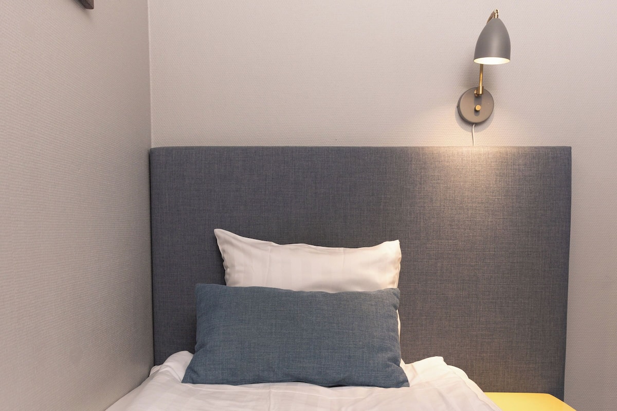 A neatly made bed with white and blue bedding, a gray headboard, and a small bedside table with a lamp.