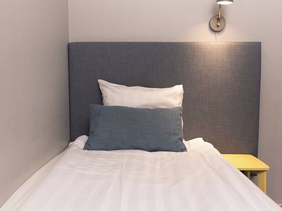 Ein ordentlich bezogenes Bett mit weißer und blauer Bettwäsche, ein grauer Kopfteil und ein kleiner Nachttisch mit einer Lampe.