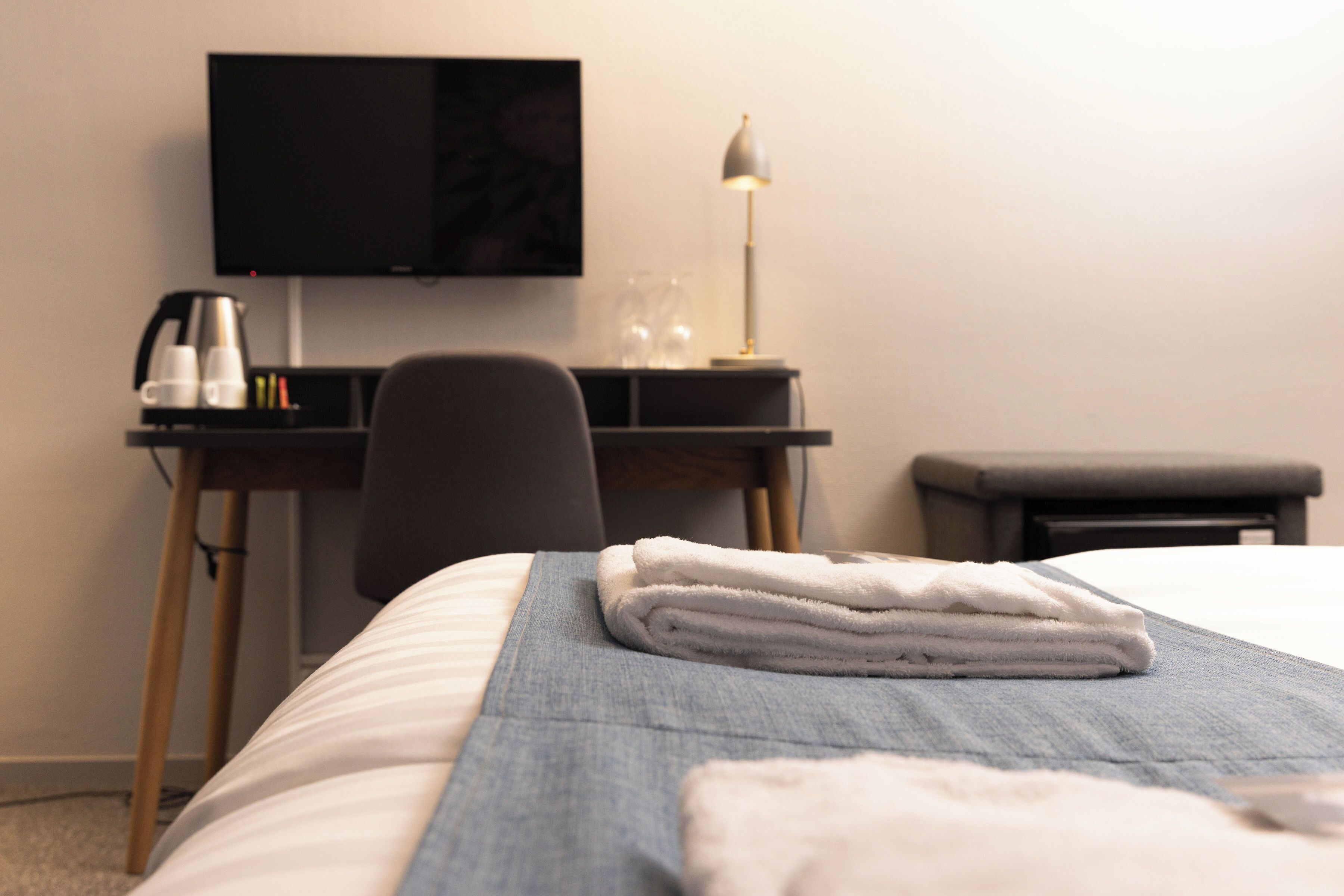 Un letto rifatto con cura, un asciugamano bianco piegato, un comodino con una lampada, una TV a schermo piatto e una macchina del caffè.