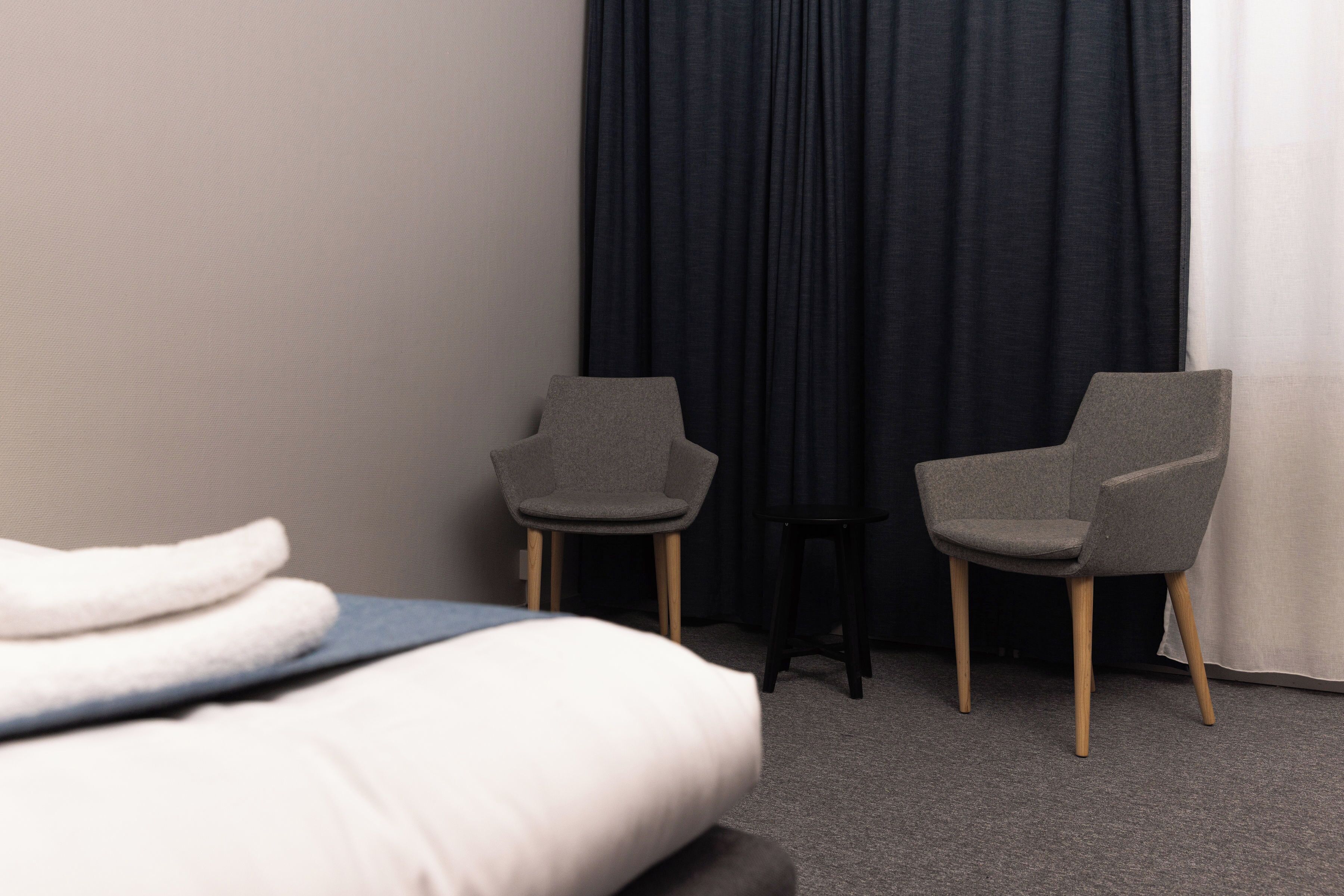Une chambre d’hôtel avec un lit, deux fauteuils gris, un petit tabouret et un rideau foncé.