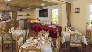A dining area with wooden tables and chairs, a kitchen in the background, and a window with curtains. - Hotel Waldidyll (Ostseebad Baabe)