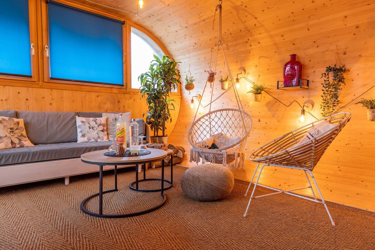 A cozy room with a wooden ceiling, a sofa, a hanging chair, and various plants.