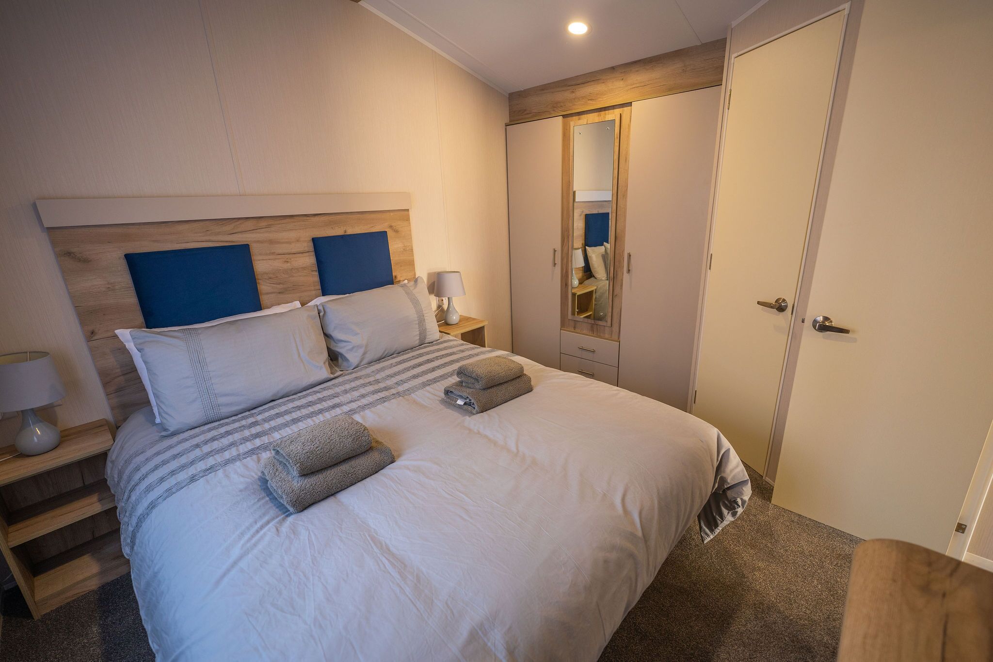 A neatly made bed with pillows and a blanket, a bedside table with a lamp, and a wardrobe with a mirror.