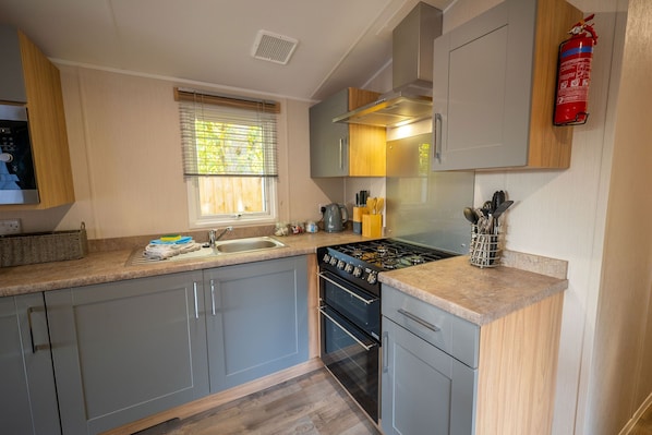 A compact kitchen with grey cabinets, a gas stove, a sink, and a window with blinds. - The Travellers Rest (Halesworth)