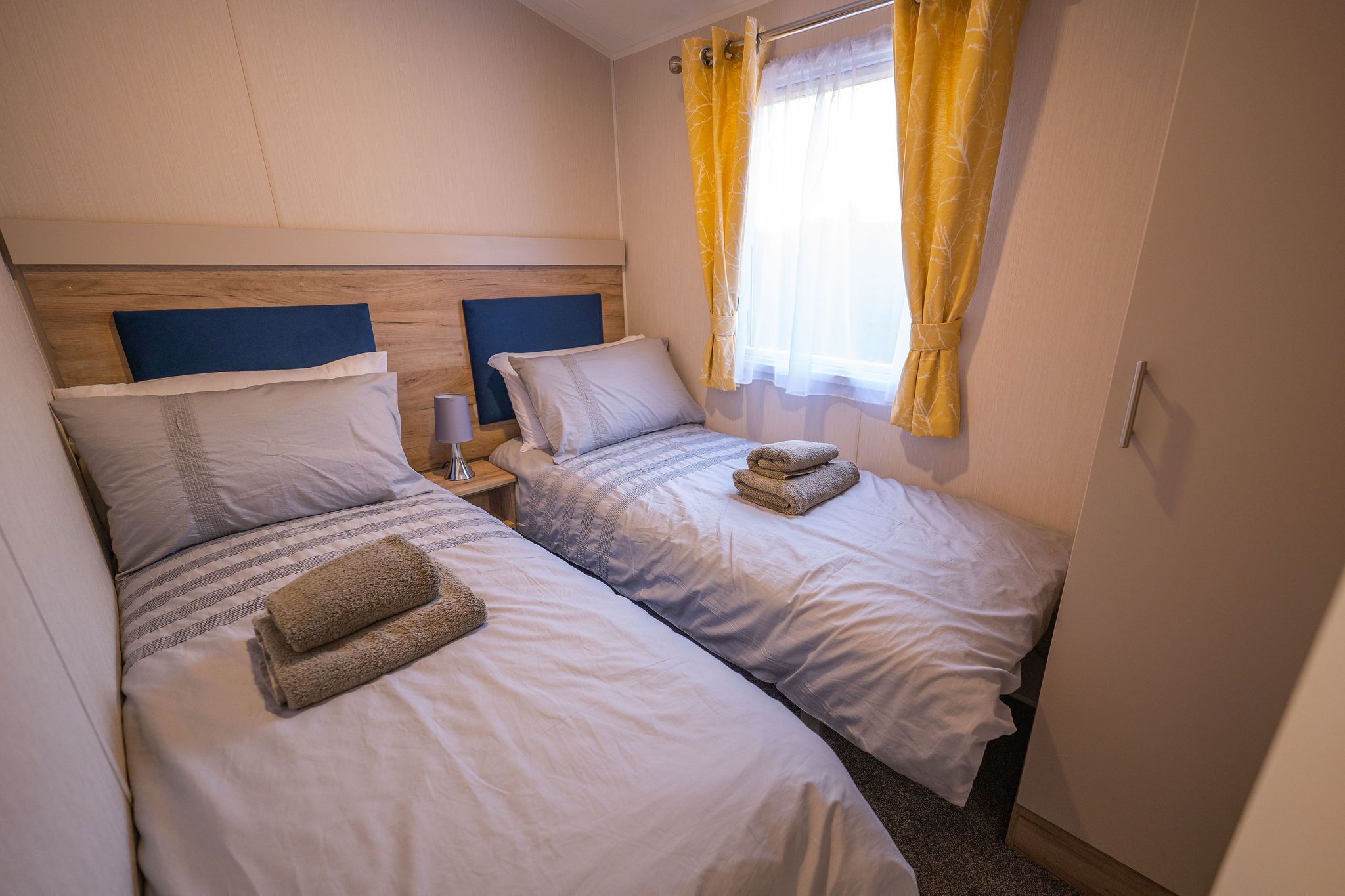 Ett hotellrum med två separata sängar, en sänggavel i trä, ett nattduksbord med en lampa och ett fönster med gula gardiner.