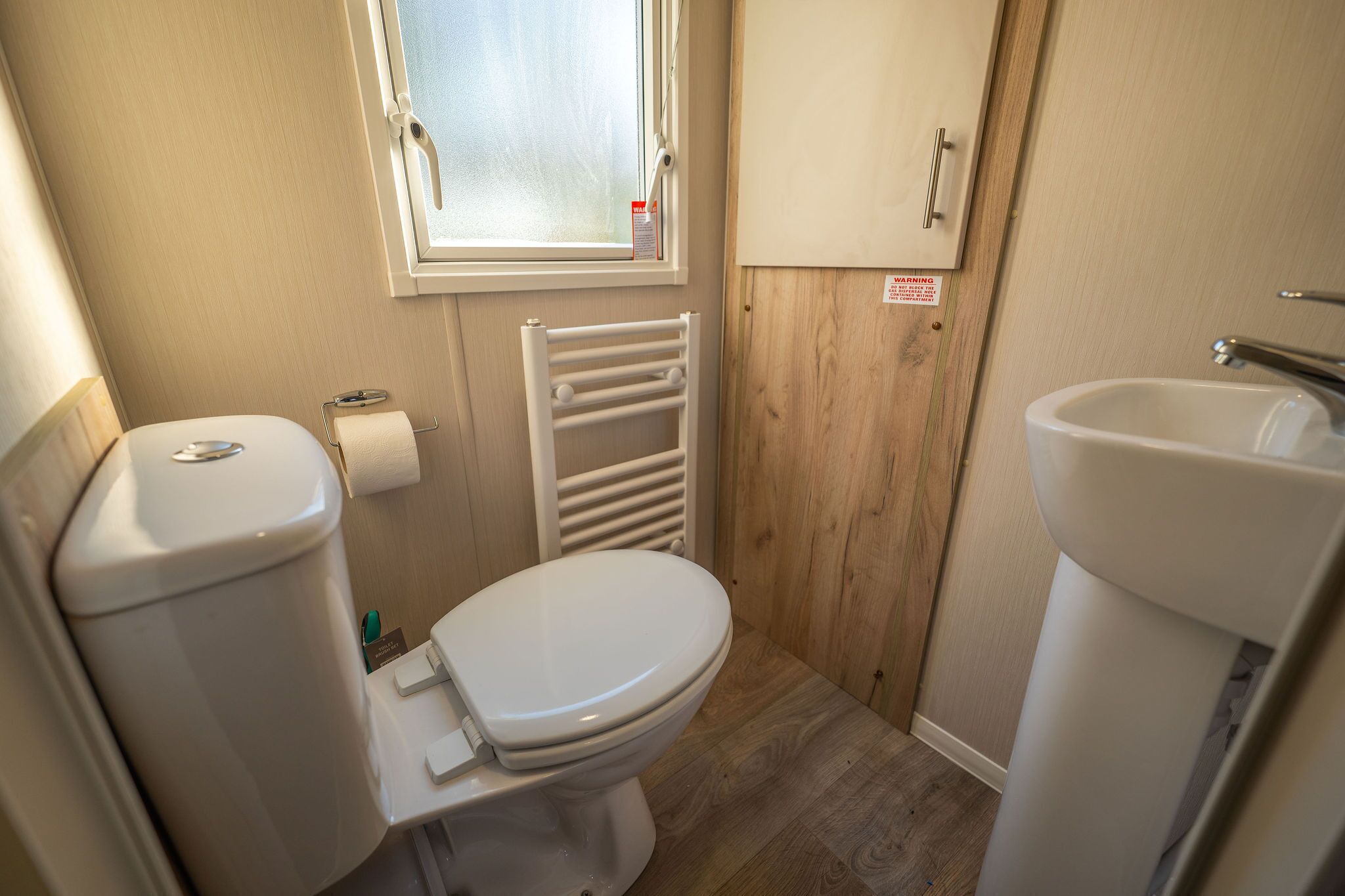 A compact bathroom with a toilet, a sink, a window, and a wooden wall.