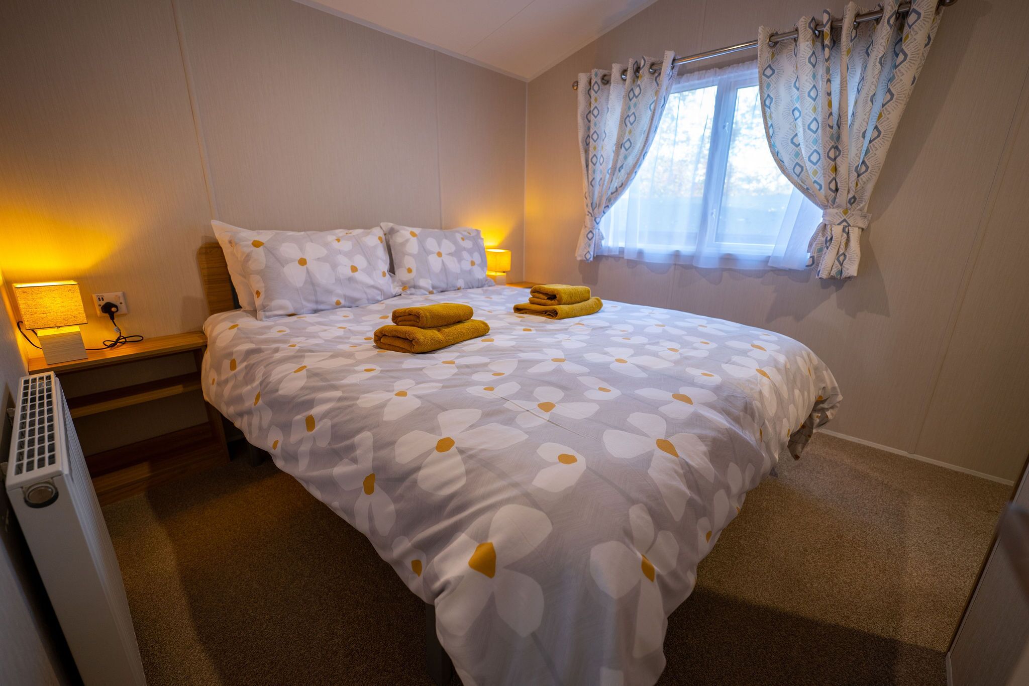 A neatly made bed with a floral bedspread and two folded towels, a bedside lamp, and a window with patterned curtains.
