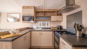 A compact kitchen with a microwave, stove, and sink. There are wooden cabinets and a window above the sink. - The Travellers Rest (Halesworth)