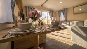 A dining area with a wooden table set for two, a vase with flowers, and a sofa in the background. - The Travellers Rest (Halesworth)