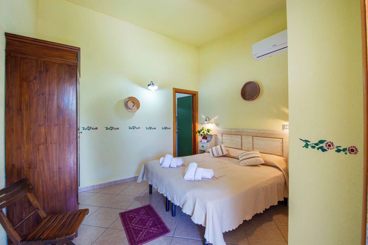 A bedroom with a wooden door, a bed with towels, a nightstand, and a wall-mounted air conditioner.