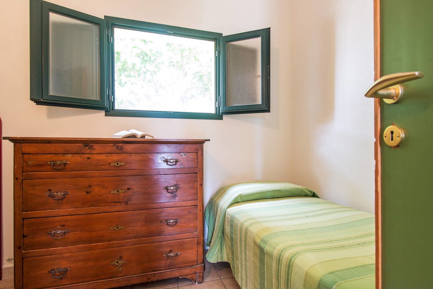 A bedroom with a wooden dresser, a bed with green bedding, a window with a view of trees, and a green door with a lock.