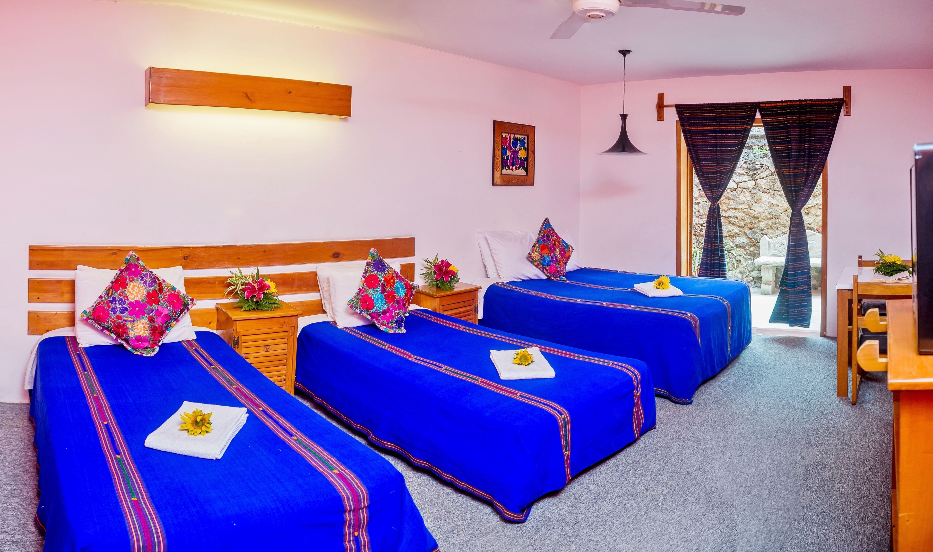 Habitación de hotel con cuatro camas, cada una con ropa de cama azul y almohadas de colores. Se observa un cabecero de madera, una mesita de noche de madera con una flor, y un ventilador de techo. Se ve una ventana con cortinas y una pared de piedra en el exterior.