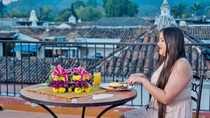 A woman sitting at a table with a meal and flowers, overlooking a cityscape with traditional buildings and a dome structure. - Hotel Panchoy (Antigua Guatemala)