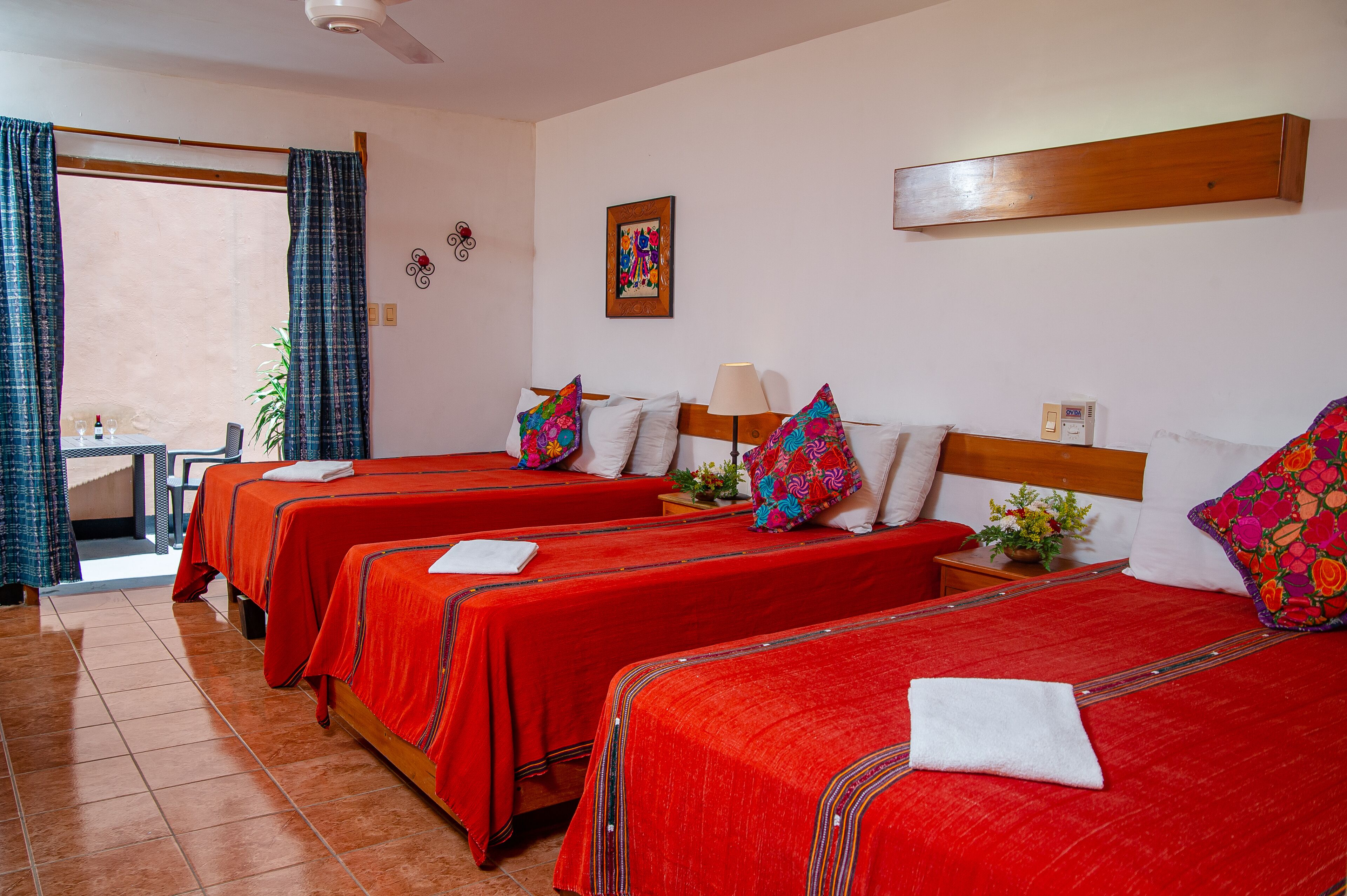 Camera d'albergo con quattro letti, ognuno con biancheria rossa e cuscini colorati. C'è un tavolino con una lampada e una pianta in vaso.