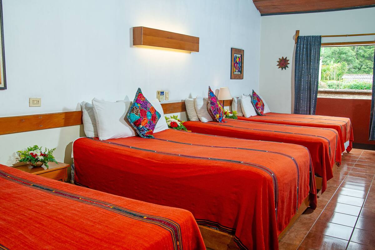 Une chambre d’hôtel avec quatre lits, chacun recouvert d’une couverture rouge et doté d’une tête de lit en bois. Il y a une lampe fixée au mur au-dessus des lits, une fenêtre avec des rideaux et un tableau accroché au mur.