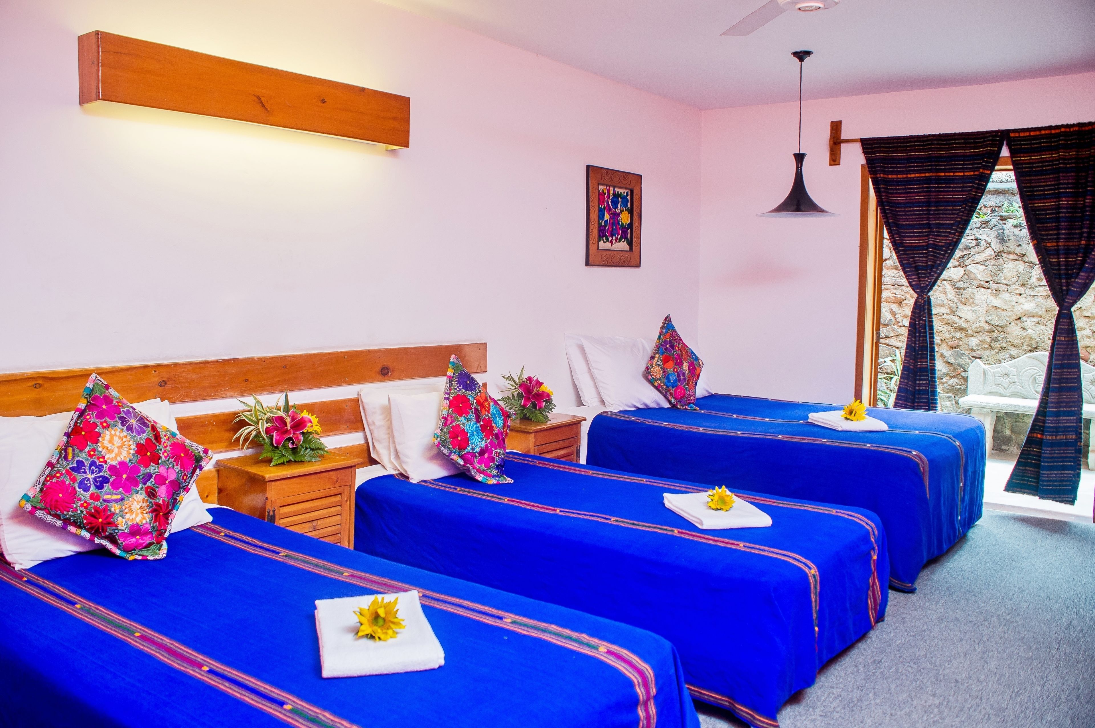 A hotel room with three beds, each with a blue bedspread and a floral pillow. There is a wooden headboard, a nightstand with a vase of flowers, and a window with curtains.