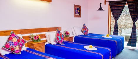 A hotel room with three beds, each with a blue bedspread and a floral pillow. There is a wooden headboard, a nightstand with a vase of flowers, and a window with curtains.