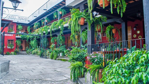 A hotel with a red facade, multiple balconies, and extensive greenery including hanging plants and potted plants. - Hotel Panchoy (Antigua Guatemala)