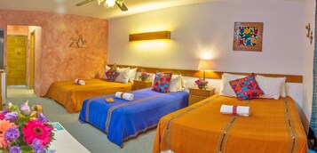 Habitación de hotel con tres camas, cada una con un edredón de color diferente, un cabecero de madera, una lámpara de pared y un cuadro con motivos florales.