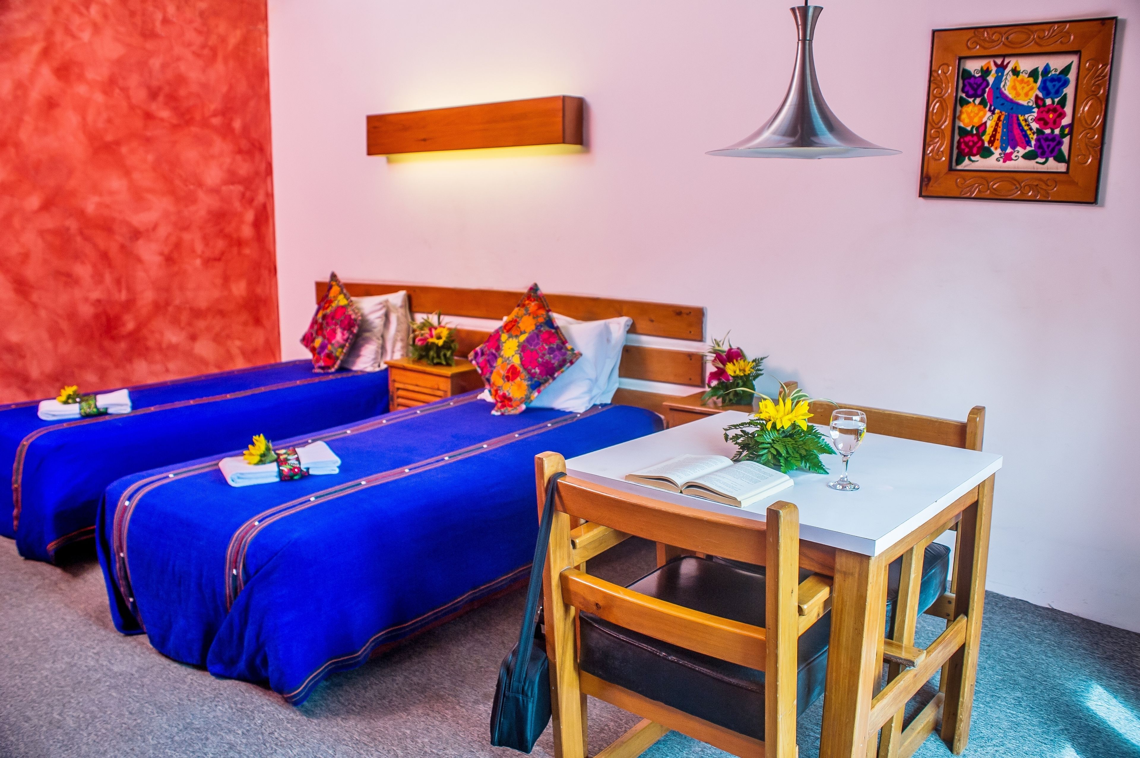 Habitación de hotel con dos camas, una mesa de comedor y una silla. Se aprecia una mesita de noche con una lámpara y un cuadro en la pared.