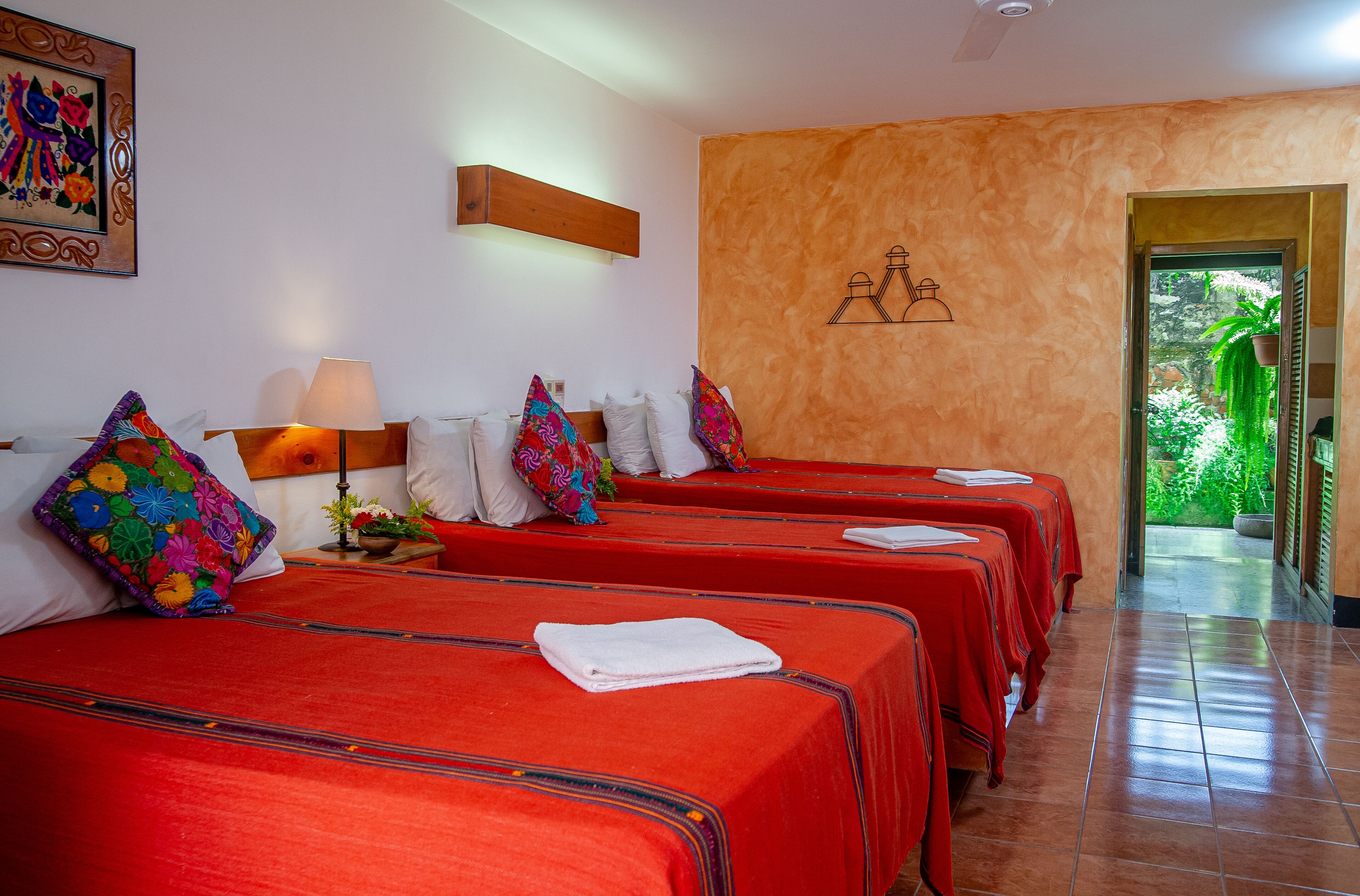 Habitación de hotel con cuatro camas, cada una con una colcha roja y almohadas de colores. Se observa un cabecero de madera, una lámpara de pared y un cuadro enmarcado sobre las camas.