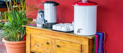 Una estación de café con cafetera, un termo y tazas sobre una mesa de madera frente a una pared roja.