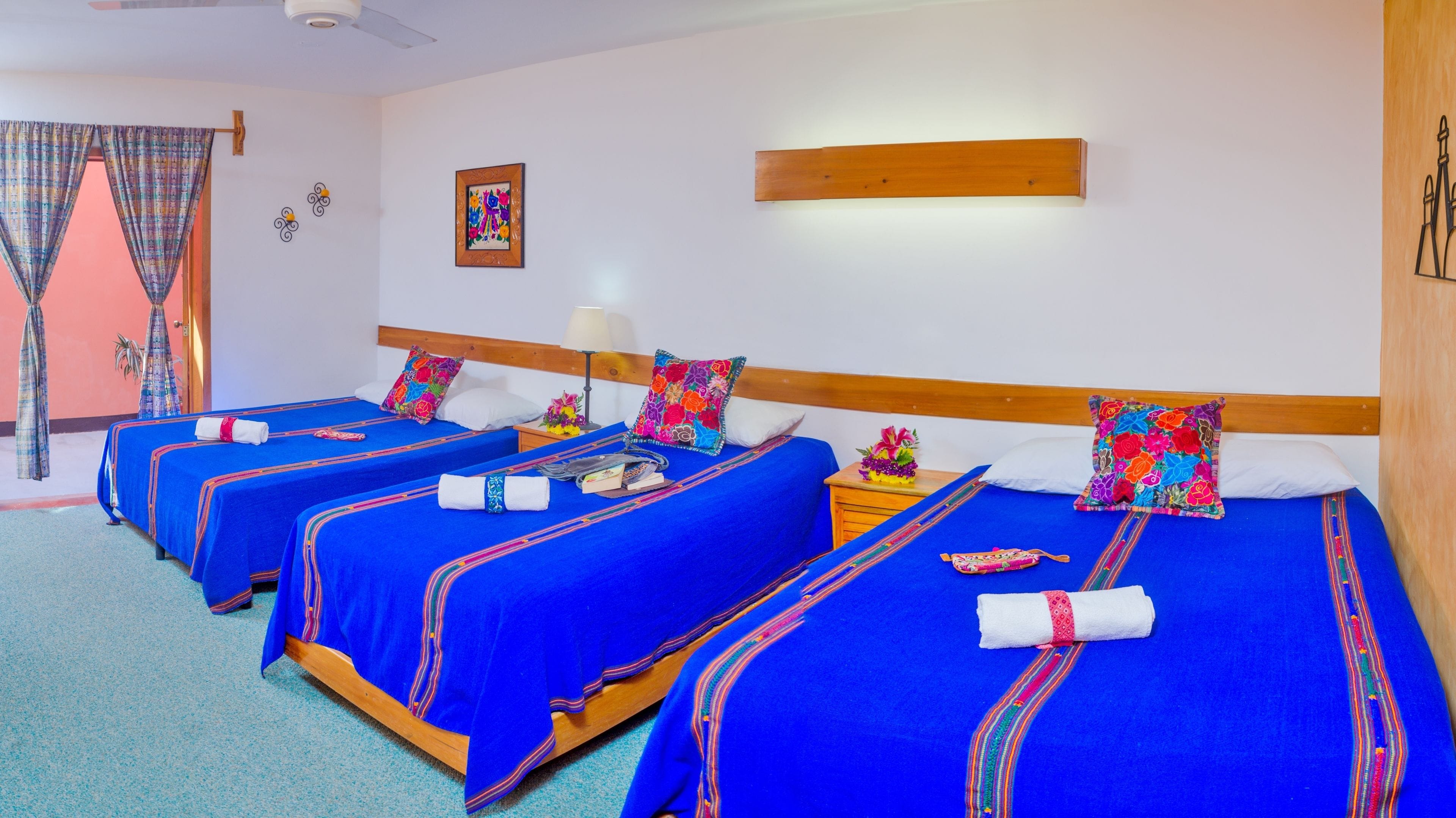 Habitación de hotel con tres camas, cada una con ropa de cama azul y almohadas coloridas. Tiene un cabecero de madera, una mesita de noche con lámpara y un estante en la pared.