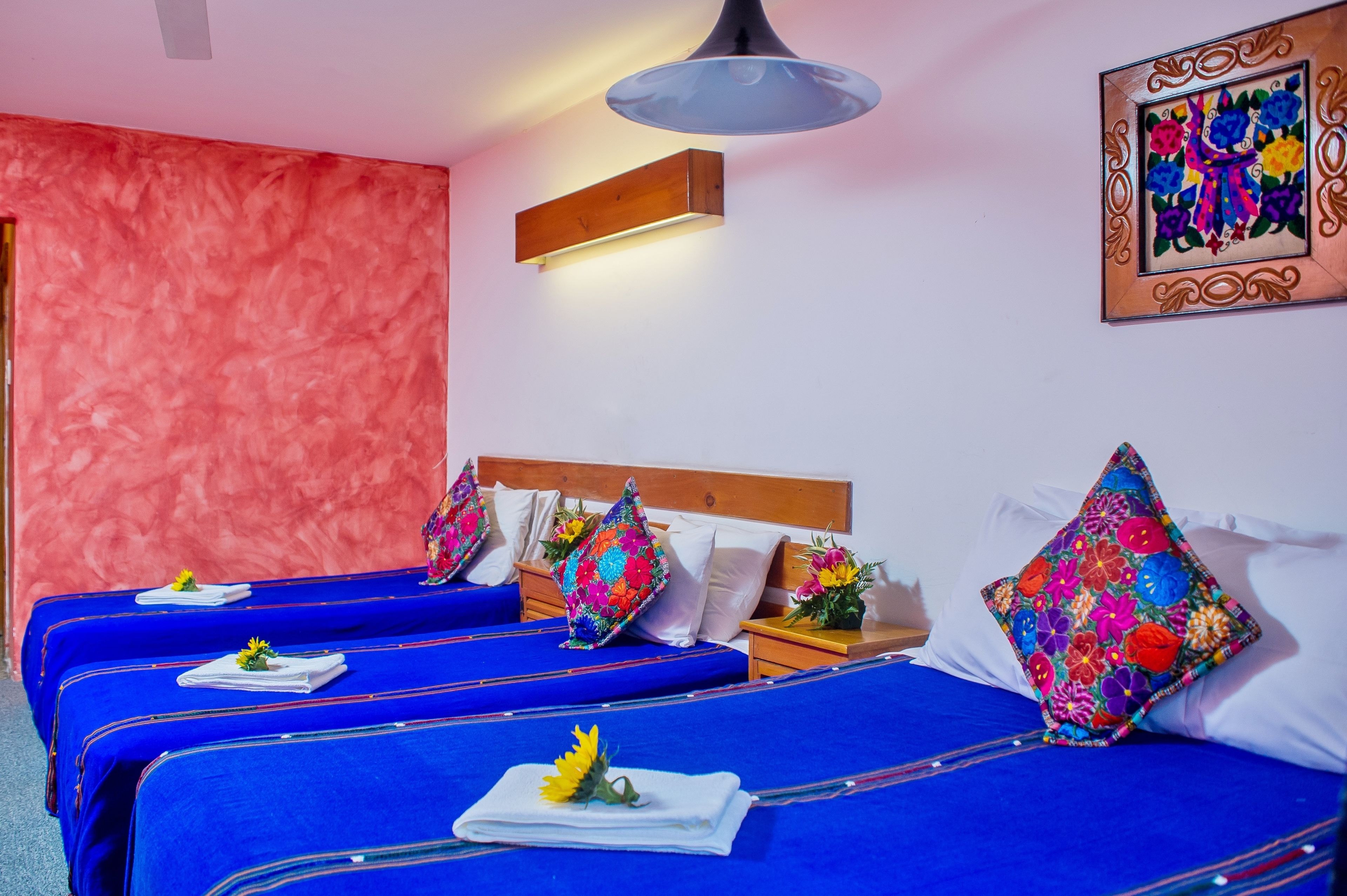 Une chambre d’hôtel avec trois lits, chacun doté de draps bleus et d’oreillers colorés. On y trouve une tête de lit en bois, un luminaire fixé au mur et une œuvre d’art florale encadrée accrochée au mur.