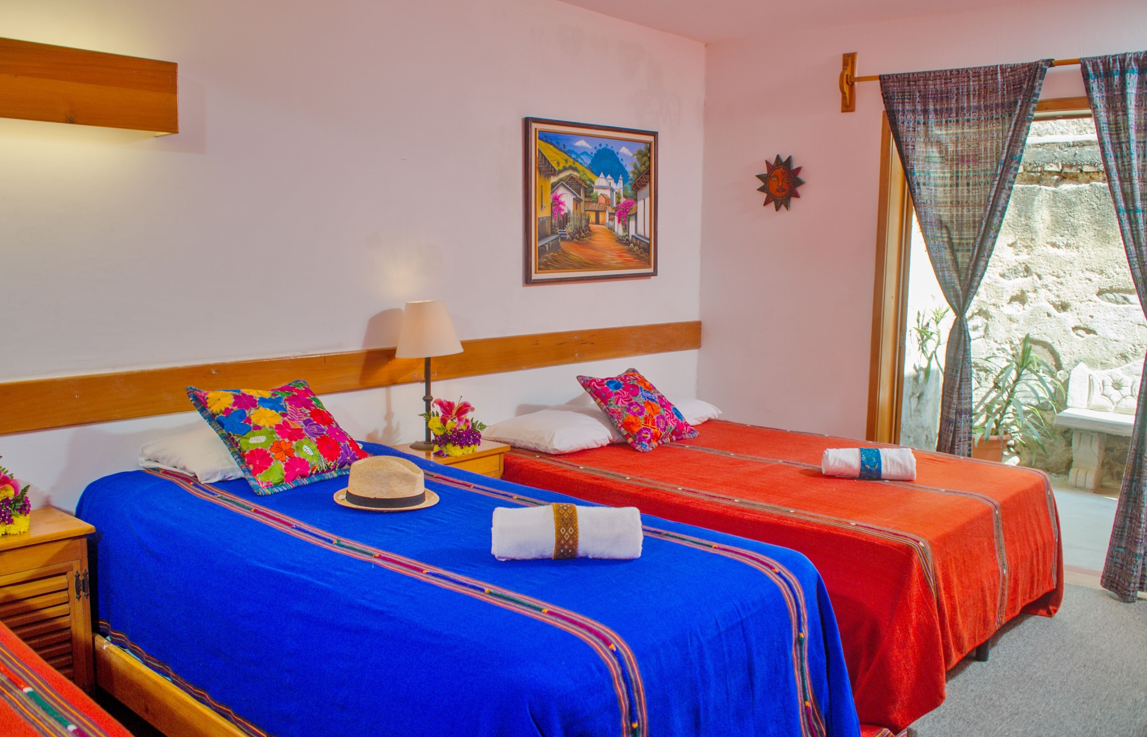 Une chambre d’hôtel avec deux lits, chacun recouvert d’une couvre-lit rouge et bleue, dotée d’une tête de lit en bois, d’une table de chevet avec une lampe et d’un tableau accroché au mur.
