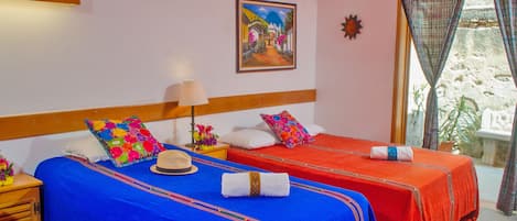 Habitación de hotel con dos camas, cada una con una colcha roja y azul, un cabecero de madera, una mesita de noche con una lámpara y un cuadro en la pared.