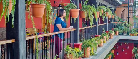 Un balcón con macetas de terracota colgantes y plantas en macetas, barandal de madera y pared roja con un mural.
