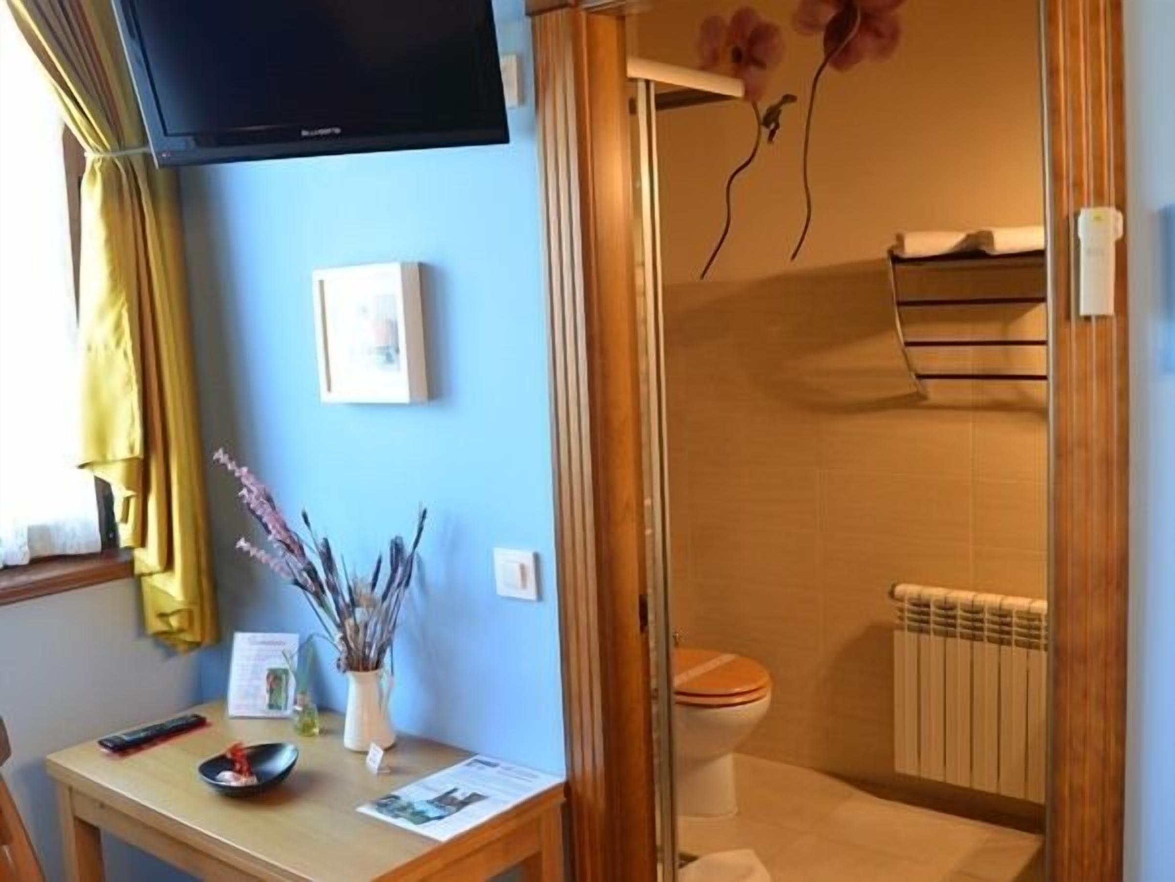 A bathroom with a toilet, shower, and a towel rack. There is a table with a remote control, a bowl, and a vase with dried plants. A television is mounted on the wall.