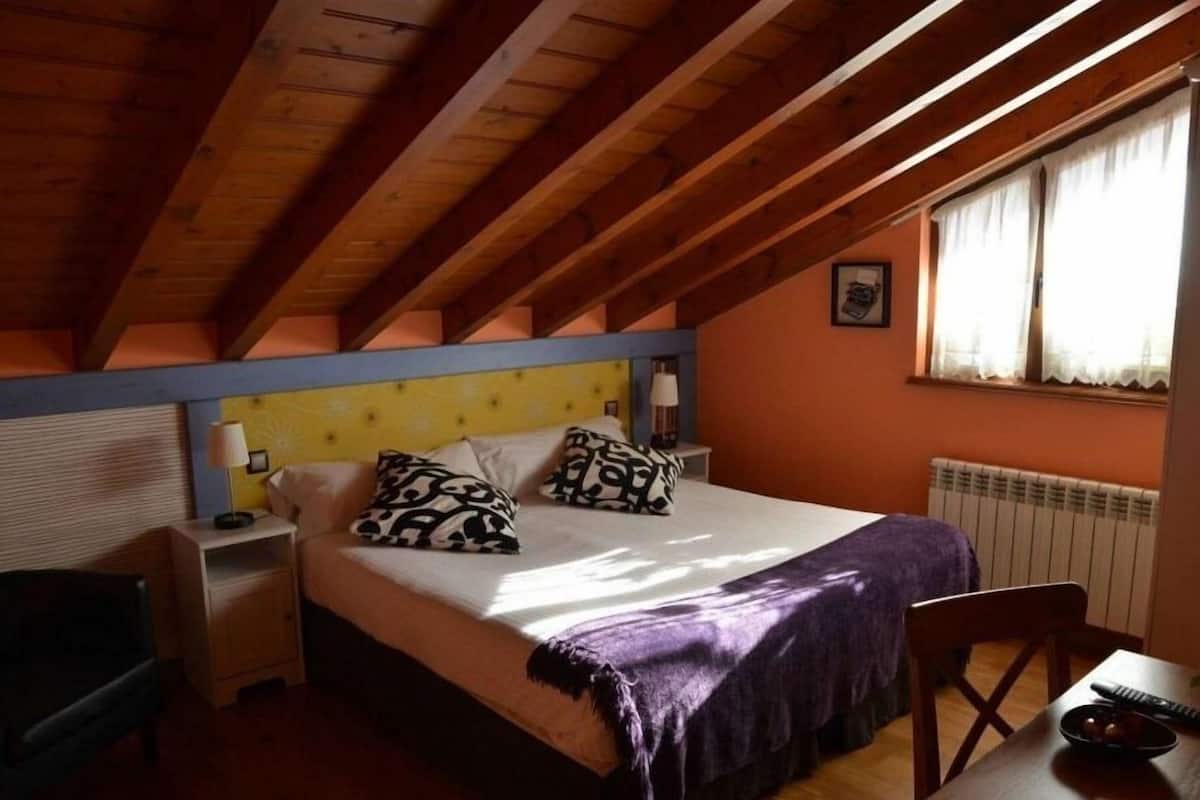 Une chambre avec un plafond en bois, un lit agrémenté d’oreillers à motifs, une table de chevet, une chaise et une table sur laquelle se trouve un bol.